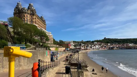 A coastal promenade next to a sandy beach on a bright day with clear blue skies. On the left side of the scene, an ornate multi‑domed historic building sits on a hillside, partly surrounded by trees. Below it runs a paved walkway with railings, a yellow public viewing telescope, and an orange life ring mounted on the railing. Along the promenade are various small shopfronts or kiosks.