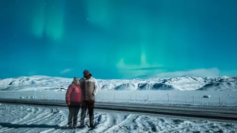 That Travel Couple Hannah Bird and Charlie Camper wearing winter coats and hats, standing with their arms around each other. They are in a snowy mountainous landscape and the dark sky is lit up with green Northern Lights.