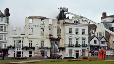 The Royal Clarence Hotel is seen partially burnt down with fire crews spraying water over it from the ground and a white crane. 