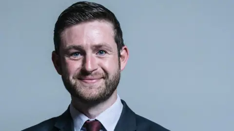House of Commons Parliamentary portrait of Jim McMahon, wearing a suit and tie.