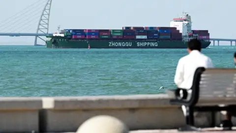 AFP via Getty Images A person sits on a bench next to the sea. In the distance a container ship heads towards a port in Kuwait City.