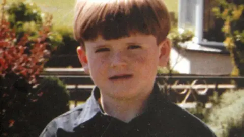 Family photo Eight-year-old Oran Doherty wearing a black shirt with an open collar.  He has short, reddish blonde hair styled in a bowl cut