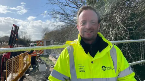 Image shows Stephen Hardy standing slightly to the right of the camera. He is wearing a bright yellow florescent jacket, which says Canal and River Trust on it on the right hand side. On the left hand side of the jacket it says Work Together - Work Safely. Underneath his jacket he is wearing a black fleece. Behind him are some trees on the right, but on the left is a metal yellow fence which overlooks the canal. The crane arm can be seen on the left and is chained up to the new gate. A workman wearing an orange florescent jacket, white helmet and black trousers and boots is standing behind the yellow metal gate looking down at the works below him.