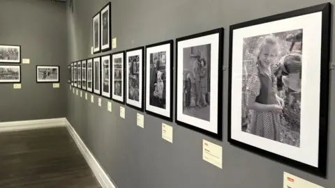 LEONARD MCCOMBE A row of photographs in black frames hanging on a grey gallery wall, The one at the front features a fair-haired child with plaits smiling.