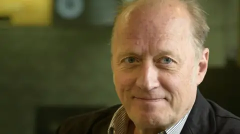 BBC/Amanda Benson A man with receding fair hair wearing a striped shirt and dark jacket.