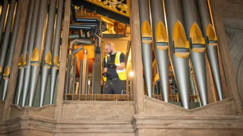 Wells Cathedral A close-up of the organ pipes with a man behind, in the process of dismantling the inner workings. He is wearing a yellow hi-bis jacket and black gloves and holds part of a pipe in his hands
