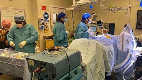 A room of medics during Sam Naylor's surgery in the RVI hospital. Four medics can be seen, three are wearing blue overalls and blue caps and masks. A nurse is to the left of the photo wearing a patterned overall and a pink cap. Sam - the patient - is on the bed completely covered by sheets. There are various pieces of equipment in the room.