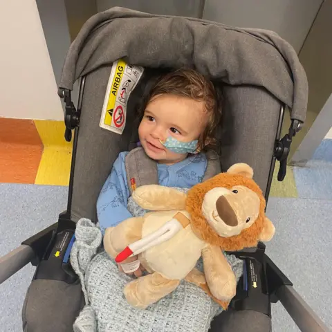 A little boy sitting in a push chair. He is looking off to the side and smiling. He has a medical tube up his nose and taped to the side of his face. There is a cuddly toy lion sat on his knee.