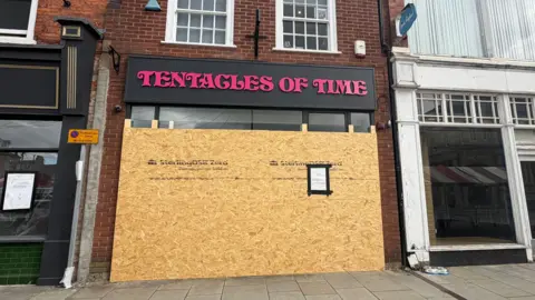 Black and pink shop front with a boarded up window
