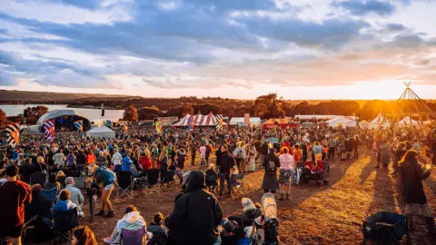 Crowds of people on a festival site, with the sun setting nearby