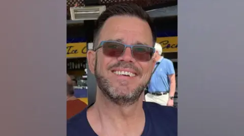 Wiltshire Police A head and shoulder image of Jonathan Neilan smiling and wearing sunglasses and a navy t-shirt. 