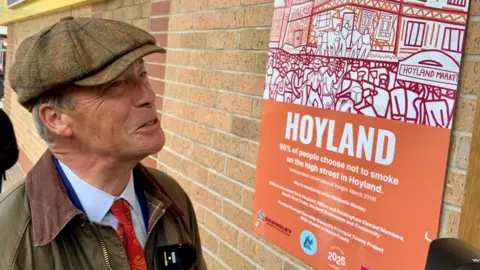 BBC/Lucy Ashton A man wearing a flat cap, a blue shirt, red tie and waxed jacket is looking at an orange sign on a wall which talks about Hoyland