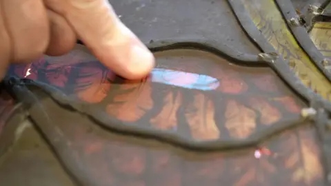 Shaun Whitmore/BBC A close-up picture of a stained glass window which is covered in soot. A man has made a line in the soot with his finger showing the layers of soot which is covering the glass. 