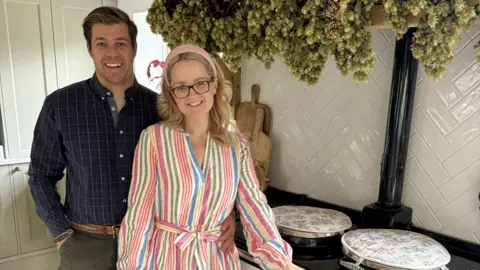 Aimee Blakey Aimee and Sean, a married couple, stand in front of an Aga unit. Aimee is wearing a striped dress in several colours and Sean is wearing a navy check shirt