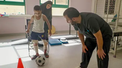 Rami kicks a football while his mother stands behind him, with a member of medical staff watching on