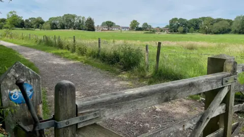A gated footpath in Pewsham, Chippenham, running adjacent to a large green field with trees and a few house in the distance. 