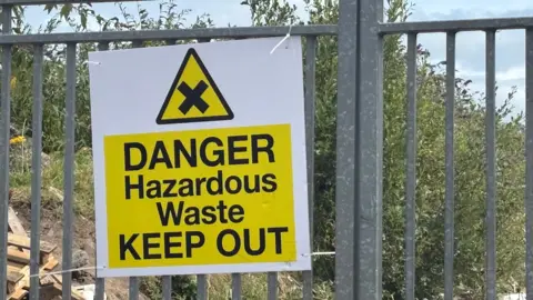 Image of sign on corrugated iron gate. The sign is yellow and in Black it reads DANGER Hazardous Waste KEEP OUT. 