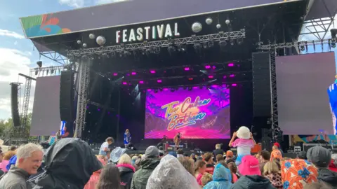 A huge crowd of people can be seen from behind, many of them wearing hoods and hats to protect themselves from the rain. Beyond them is an enormous stage with colourfully dressed performers. A large and vibrantly coloured pink billboard on the stage reads "The Cuban Brothers'.