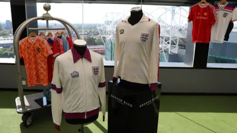 Graham Budd Auctions Viv Anderson's white England shirt and tracksuit top from his debut against Czechoslovakia in 1978 are on mannequins, while a number of other jerseys picked up during his career are on clothes hangers behind them. The items are displayed at Manchester's Hotel Football with the Old Trafford stadium partly visible in the background through the window.