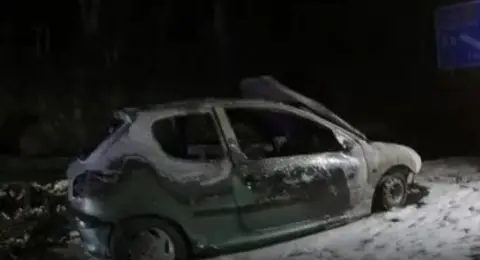 The burnt out car sitting on the side of the road. It is charred and grey.