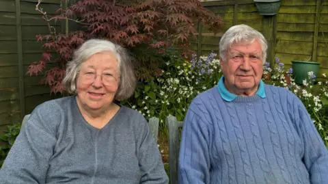 Michelle and Nigel Parry sit in their garden in front of flowering shrubs. Michelle has white/grey hair and a grey cotton top. Nigel has white hair and wears a blue jumper over a polo shirt.