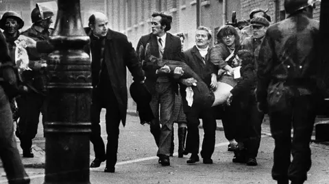 A picture taken on 30 January 1972 - Bloody Sunday. A group of men are carrying a lifeless body - they are accompanied by a priest waving a wife handkerchief. There are soldiers at the side of the picture and in the foreground.