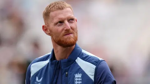 PA Media Ben Stokes looking to the right with pursed lips. He has short, ginger hair and a ginger beard and is wearing a blue England bodywarmer and matching blue t-shirt. The background is out of focus.