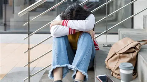 A young person with long brown hair is sitting alone on concrete steps, their head resting on folded arms over bent knees. They are wearing a white jacket with red sleeve stripes, a yellow top and light blue frayed jeans. Beside them are a tan backpack, a pink‑cased phone, and a takeaway coffee cup.