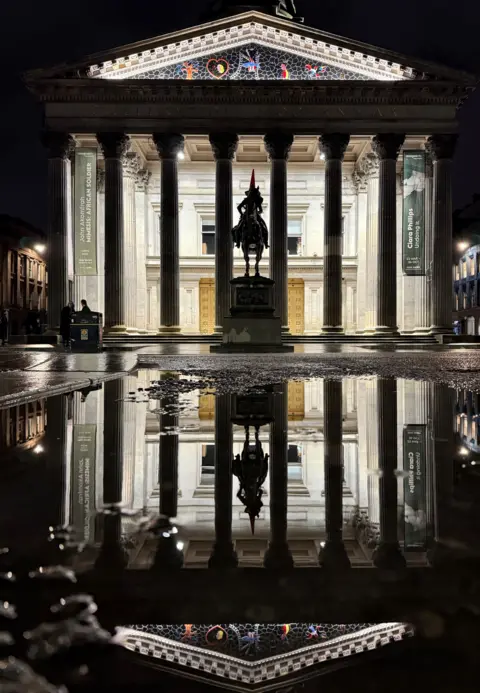Bill Cameron The Gallery of Modern Art - a neoclassical building with large columns and a statue of the Duke of Wellington on a horse - on a wet night, with the reflection of the building visible in a puddle. 