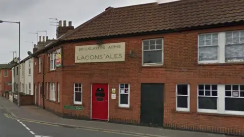 Google The exterior of a pub. It is a red brick, two-storey building on the corner of a street. It has a red painted entrance door. Above the door is a white side on the wall saying "BRICKLAYERS ARMS" AND "LACONS ALES".