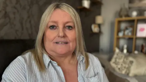 Louise has medium length blonde hair and is wearing a striped blue and white shirt. She's looking at the camera while sitting on a sofa in her living room which has white walls and a silver papered wall. Behind her over her left shoulder is a cream cushion and some shelves with a variety of items on them. The background is out of focus.