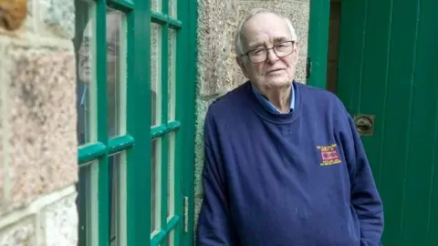 BBC An older man with grey receding hair and glasses leans against a granite wall looking towards the camera. He wears khaki, flannel trousers and a blue sweatshirt. He looks sad.