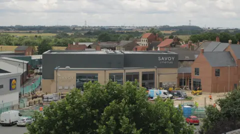 A small town scene with a modern Savoy Cinemas building under construction. Workers and vehicles are can be seen nearby. A Lidl store is on the left of the building. Countryside fields can be seen in the background under a cloudy sky. Red-brick homes surround the construction area.