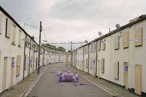 Museum Wales A group of children wearing light purple outfits all sit in at a table in the middle of a street of houses with boarded up windows and doors. There is purple bunting draped across the street multiple times and purple balloons attached to walls.