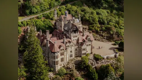 Callum Thompson/National Trust An aeriel view of the Cragside house estate. Built into the crag, the mansion has multiple chimneys poking out of its red roof. The building is grey stone and surrounded by lush trees.
