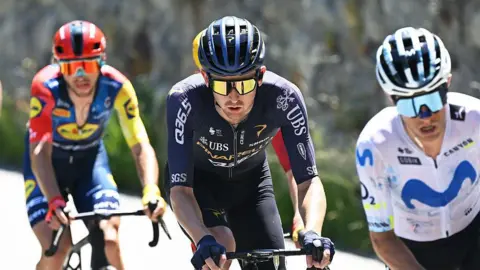 Getty Images Mark Donovan rides in the middle of two other riders in this year's Giro d'Italia. He is wearing a black-and-blue helmet and a dark purple jersey.
