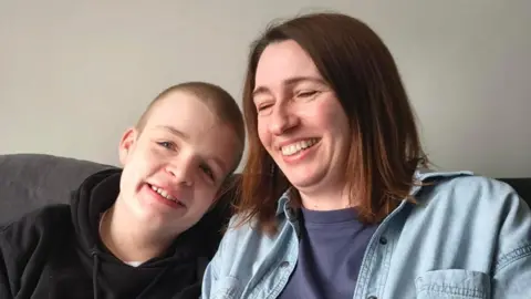 Contributed Teenager William sits on a sofa with his mum Sarah. William has a shaved head and wears a black hoodie. Sarah has short brown hair and wears a denim shirt with a navy T-shirt underneath. They are both smiling. William is looking at the camera while Sarah has her eyes shut as she laughs.