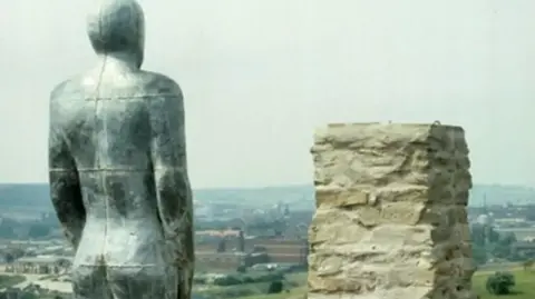 Dave Ball / Sheffield Hallam University A human-like figure, with its hands by its side, is standing next to a brick trig point. The figure overlooks a vista with buildings and fields. It is set against a pale grey-coloured sky.