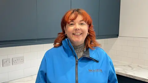 Lydia Brown is pictured smiling at the camera. She has red shoulder-length hair and is wearing a blue jacket. She is standing in a kitchen which has dark grey/blue cabinets and white marble-effect work surfaces.