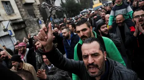 Reuters A man wearing a black jacket leads and carrying a cross leads other men in a protest. Some of them are chanting 