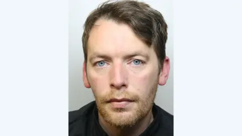 A police mugshot of Mark Manship wearing a black t-shirt and standing against a grey background. He has light blue eyes, short brown hair, and a ginger beard. 
