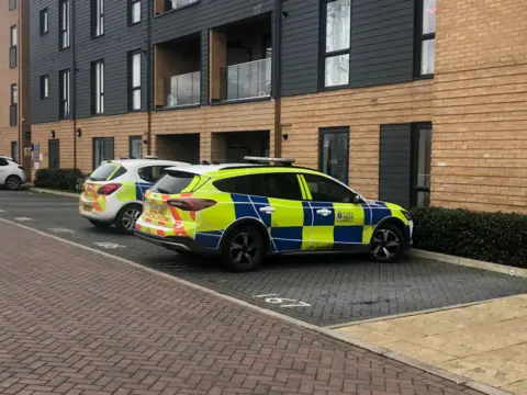 Alex Stevanovic/BBC Two police cars are parked outside a modern set of flats with balconies