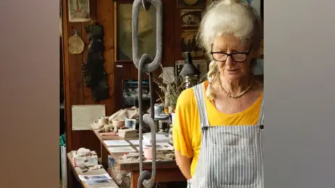 Elspeth Owen Elspeth Owen looking down in her studio, with a table and bench behind piled with ceremics