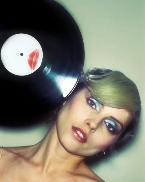 Martyn Goddard Debbie Harry poses with a vinyl record pressed to her head, featuring a lipstick kiss mark on the label, wearing blue eyeshadow and red lipstick.