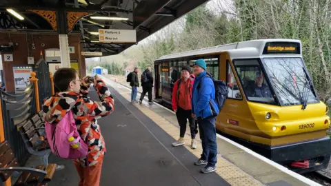 Alex Manners A man in a orange, white, beige and black jacket, with pink backpack, takes a photo of a man with red coat, glasses and black hat and a man with a blue jacket on, ginger beard and turquoise beanie hat. They are stood next to a yellow tram at a railway station 