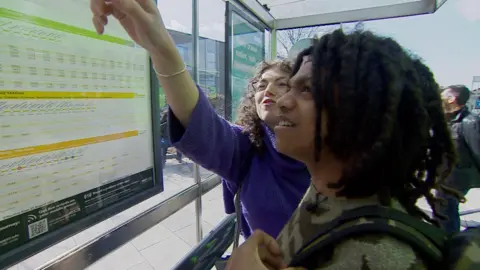A woman points to a timetable mounted in a glass bus shelter while a teenage boy watches her. It is a sunny day and people stand behind them.