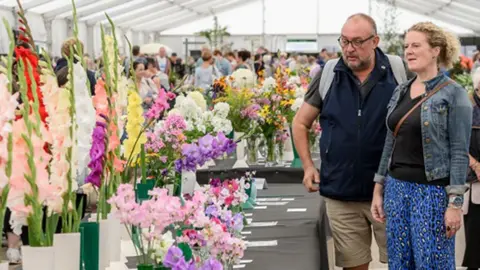 Amateur competition entries on display in the Bloomin’ Big Marquee, showing a long table of flowers, mainly gladioli, being admired by a man with glasses and a navy fleece and a woman with curly blonde hair in a denim jacket