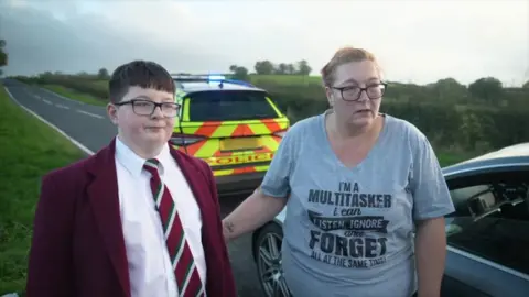 Dylan wearing a maroon blazer, white shirt, maroon/green and white school tie and black glasses standing in front of a police car with his mum who's wearing a grey top and black glasses with her hair tied up 