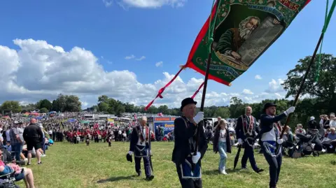 Members of the Royal Black Institution dressed in black suits and sashes hold aloft a banner while bands come behind them. Thousands of spectators can be seen