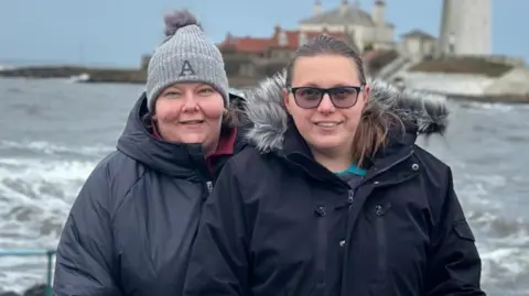 Contributed Two women stand together and smile at the camera in front of rough sea on a grey day. They both wear black coats. One woman wears a beanie hat and the other has dark hair and sunglasses. Some buildings can be seen behind them.